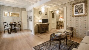 A room with a wooden floor, a dining table with chairs, a TV, a lamp, and a painting on the wall.