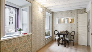A room with floral wallpaper, a wooden dining table with chairs, a window with a view of a building, and a door.