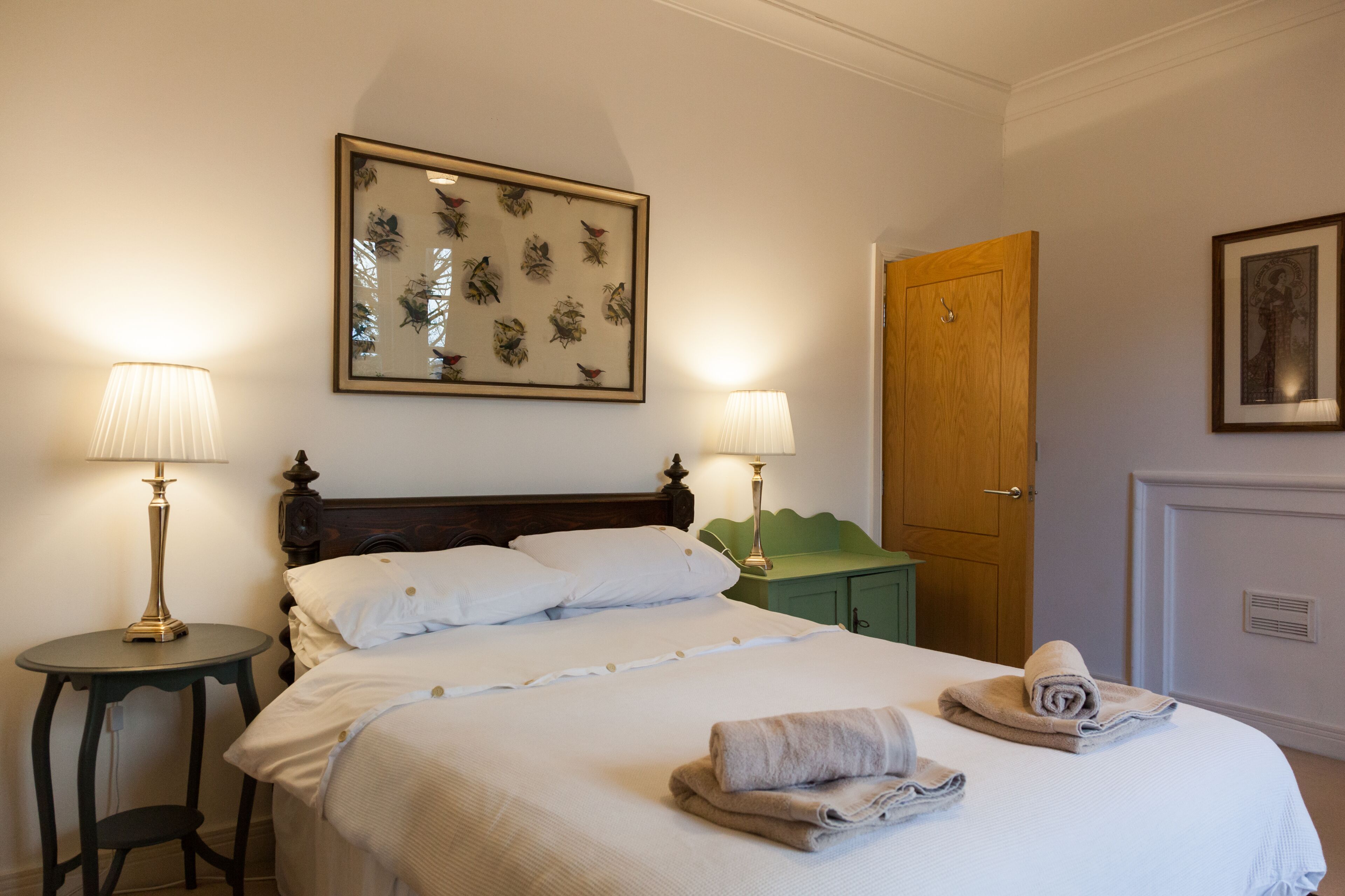 A neatly made bed with white linens, two bedside lamps, a framed floral artwork above the bed, a wooden door, and a green nightstand.