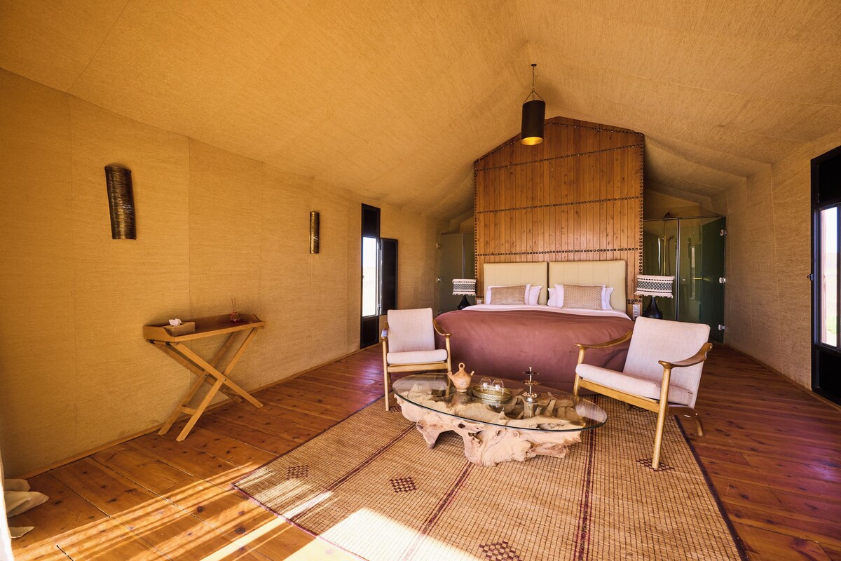 Um quarto espaçoso com uma cama grande, duas cadeiras, uma mesinha e teto de madeira.