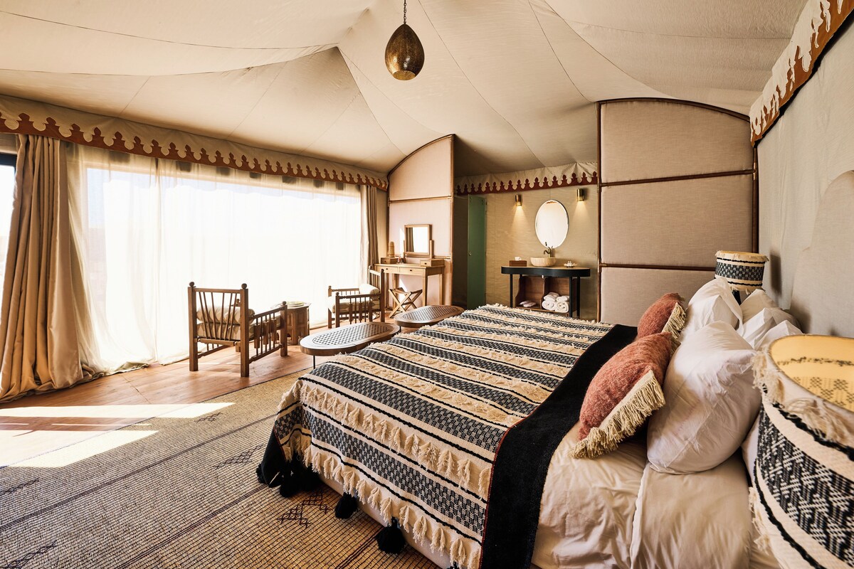 Quarto com cama grande, área de estar com cadeiras, toucador com espelho e luminária pendente.