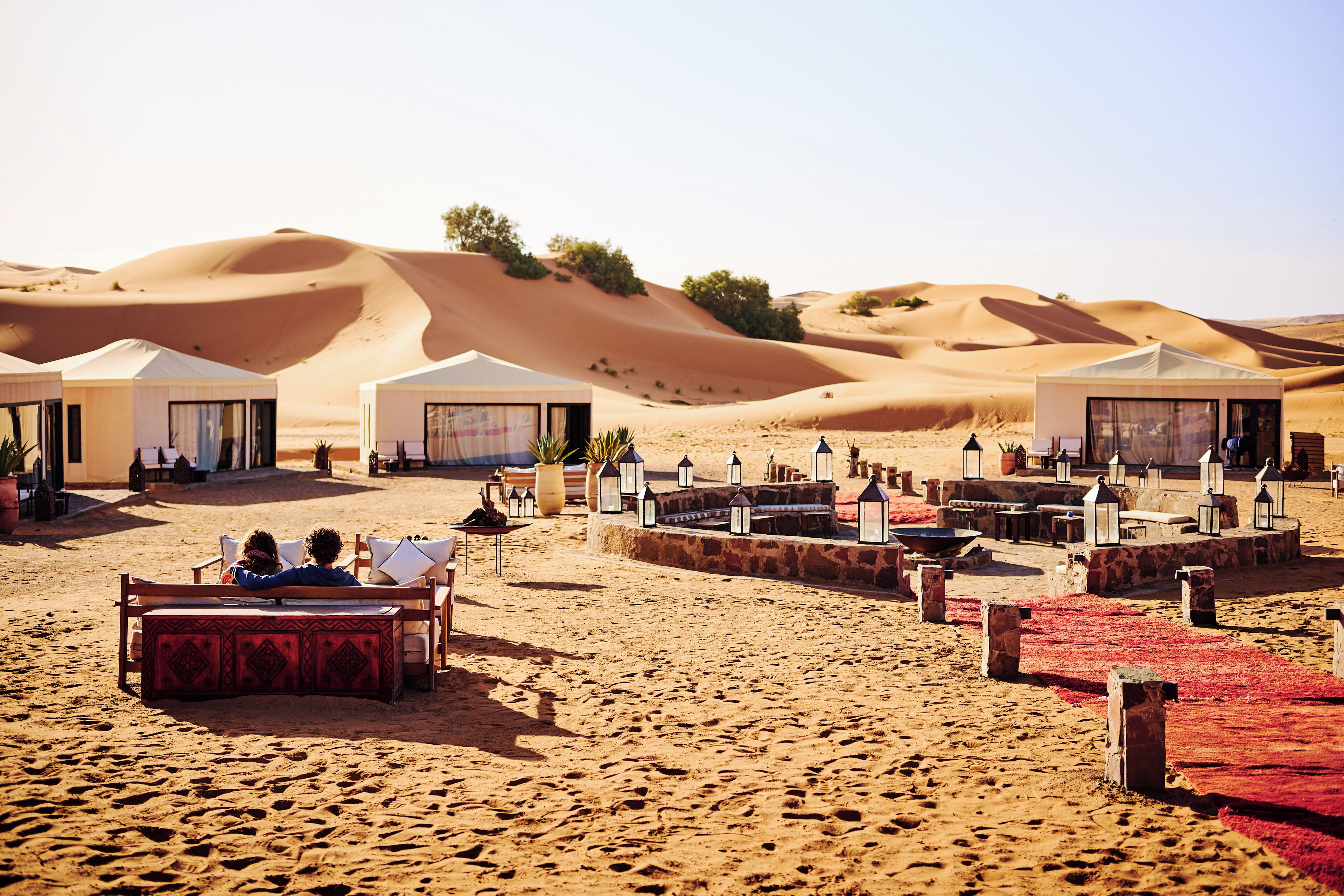 Un campamento en el desierto con tiendas de campaña, mobiliario exterior y un paisaje arenoso.
