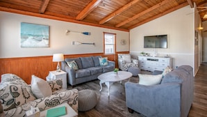 A spacious living room with a wooden ceiling, a grey sofa, a white coffee table, and a TV on the wall. - 5377- Memorial Blvd. - Family-Friendly KDH Retreat Near the Beach (Kill Devil Hills)