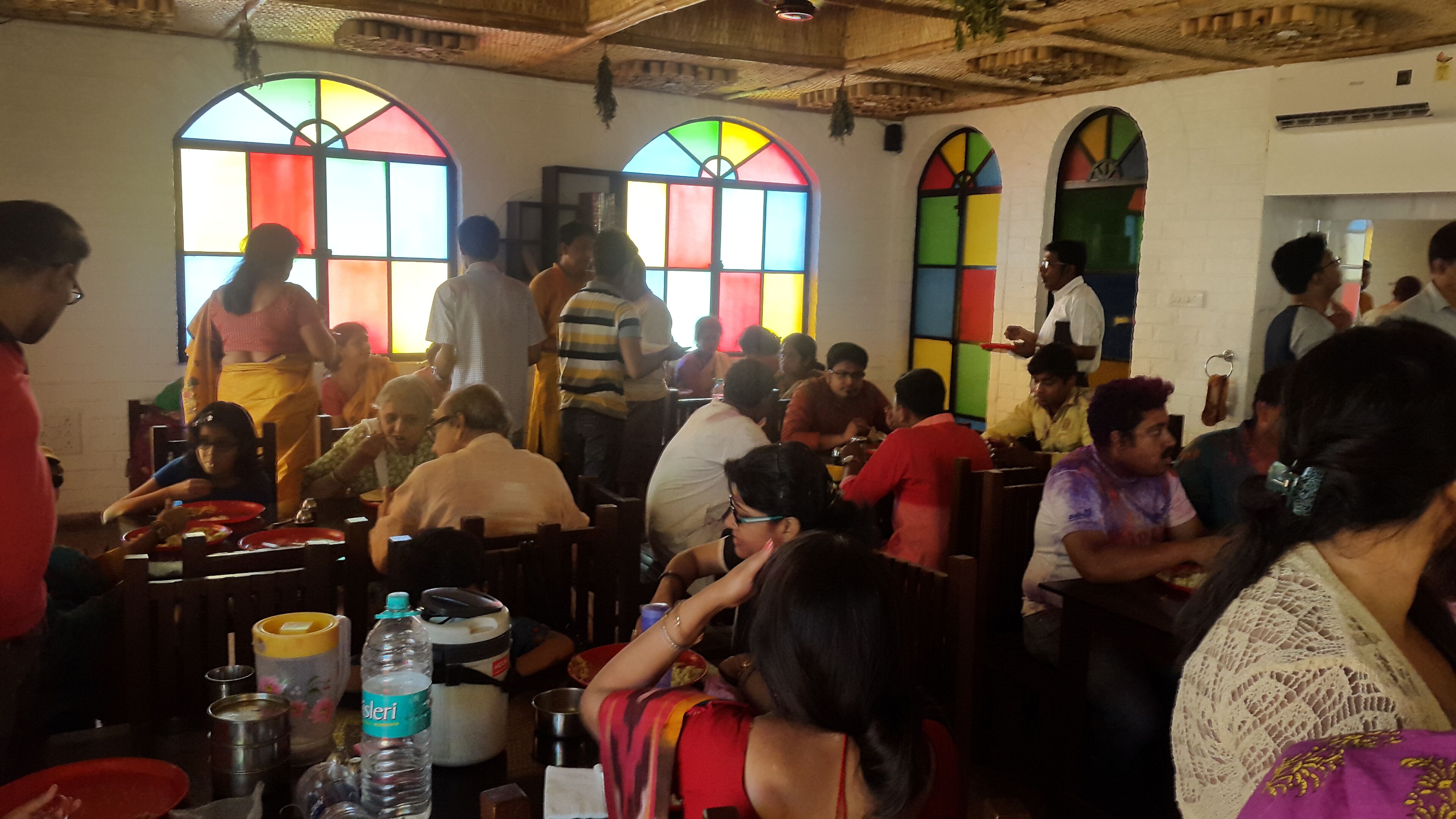 A crowded indoor space with people seated at tables, large windows with colorful stained glass, and a rustic ceiling.