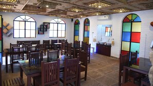 A dining area with wooden tables and chairs, large windows with stained glass, and a kitchen area in the background.