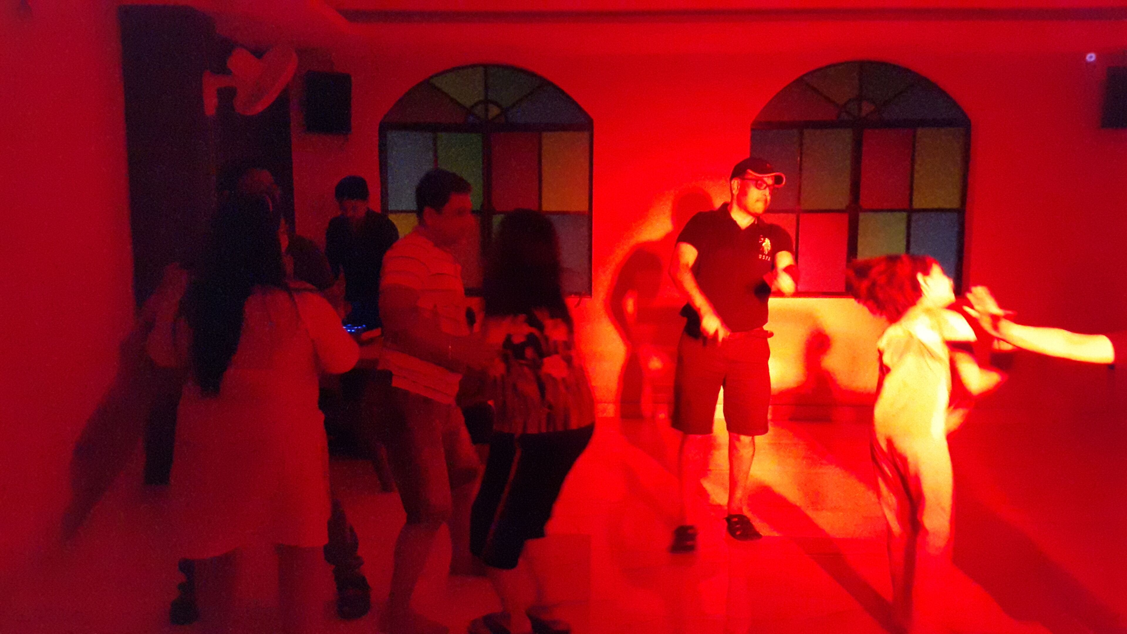 A group of people dancing in a room with arched windows and a red hue.