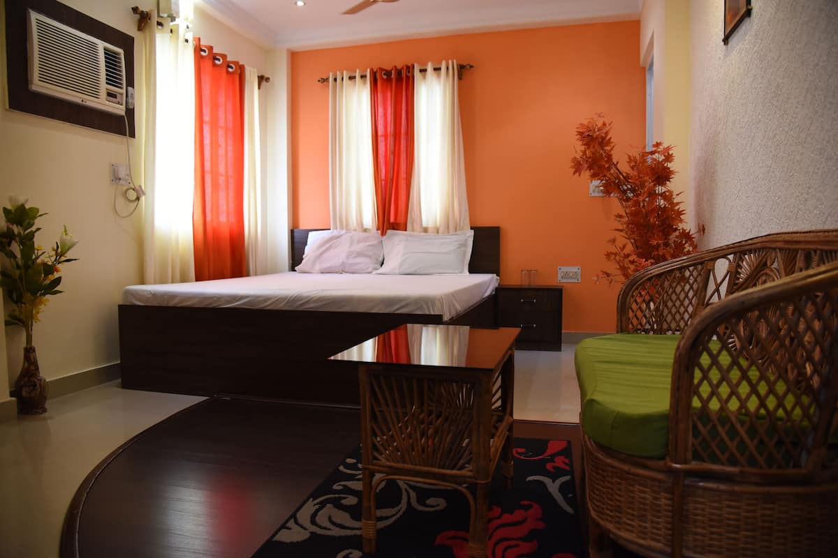 Habitación de hotel con cama, una mesita, sillas de mimbre, una planta en maceta y un equipo de aire acondicionado en la pared.