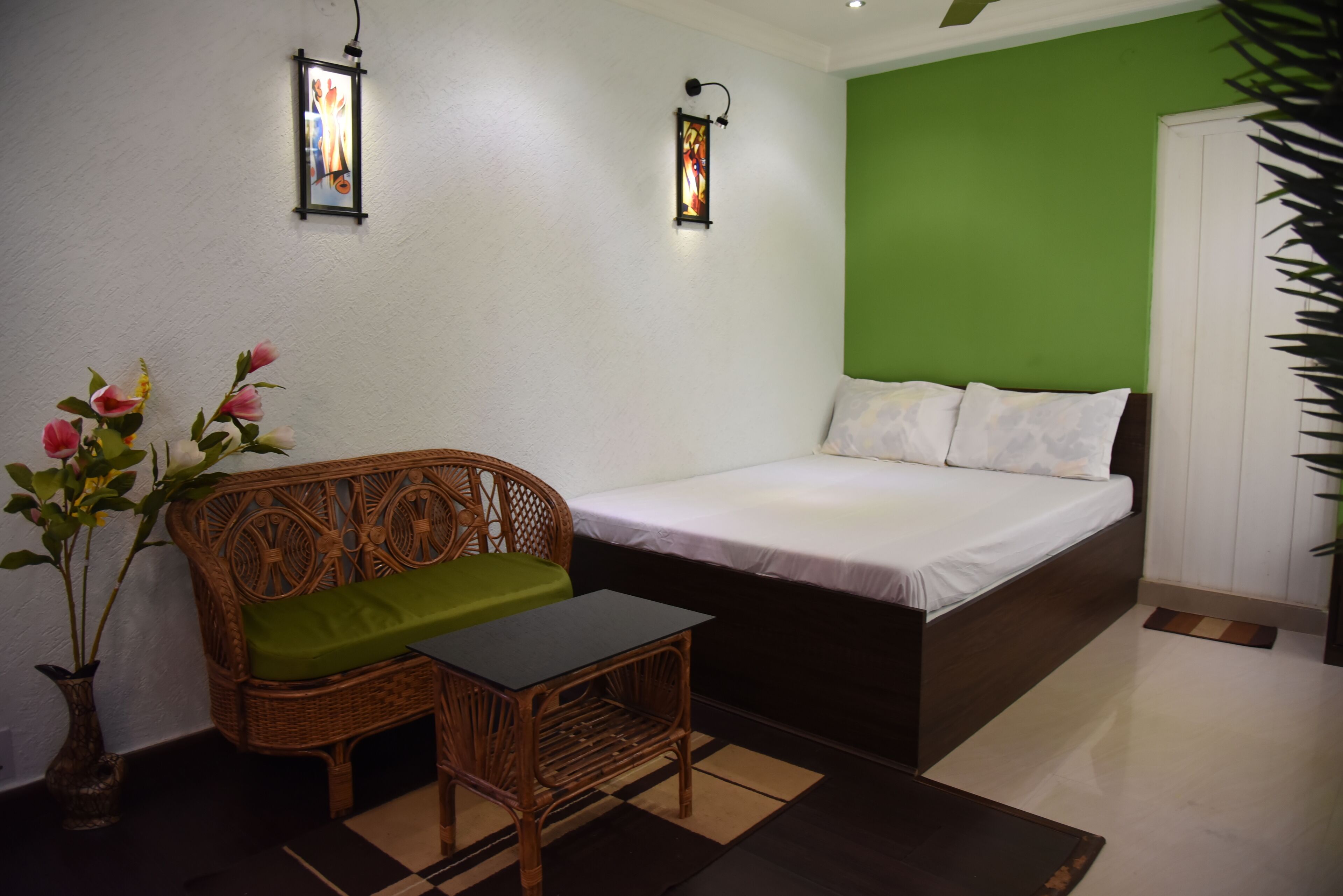 A room with a bed, a wicker sofa with a green cushion, a small table, and two wall-mounted lamps.