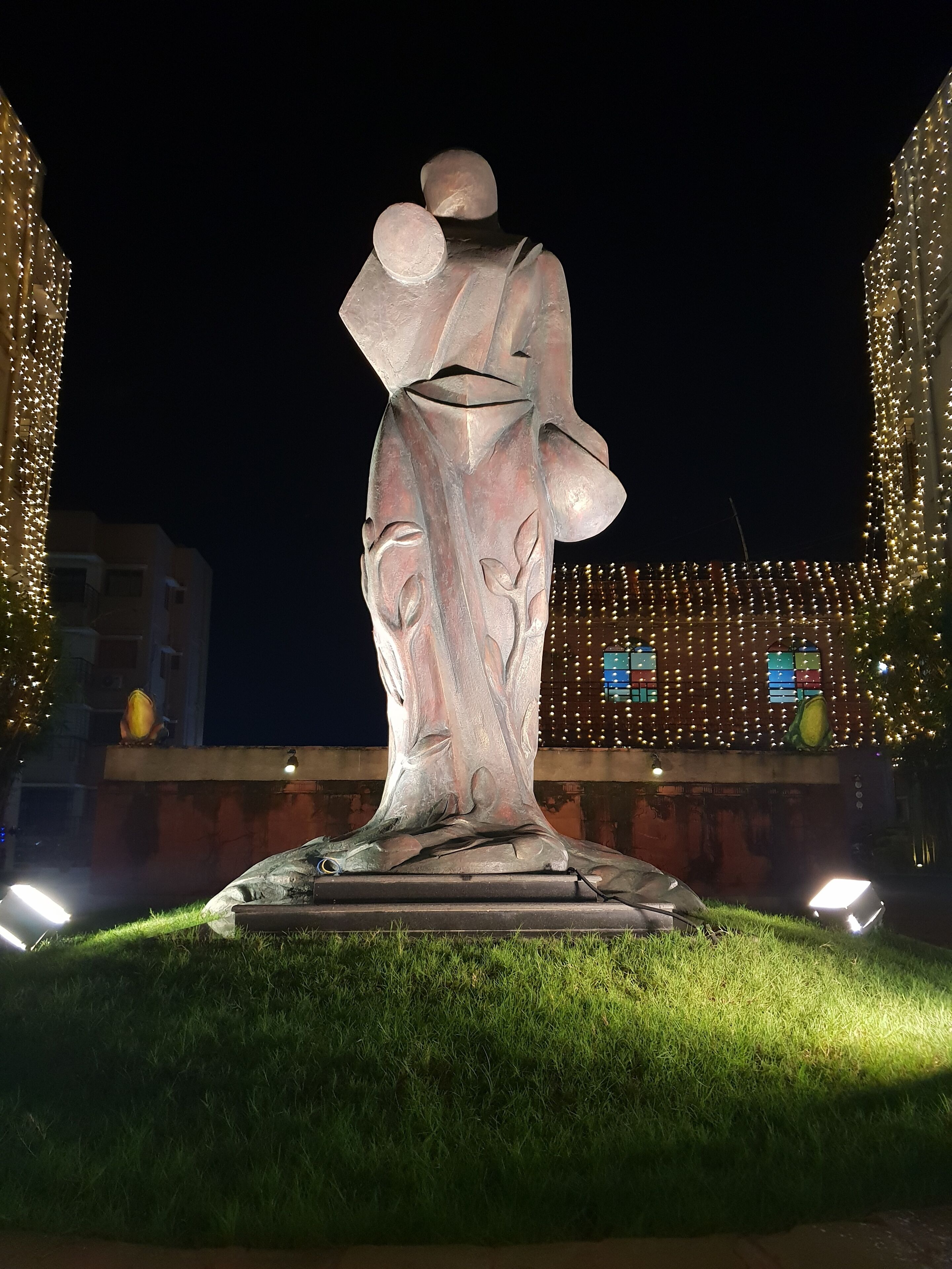 A sculpture of a figure in a flowing dress, set on a pedestal in a grassy area with lights in the background.