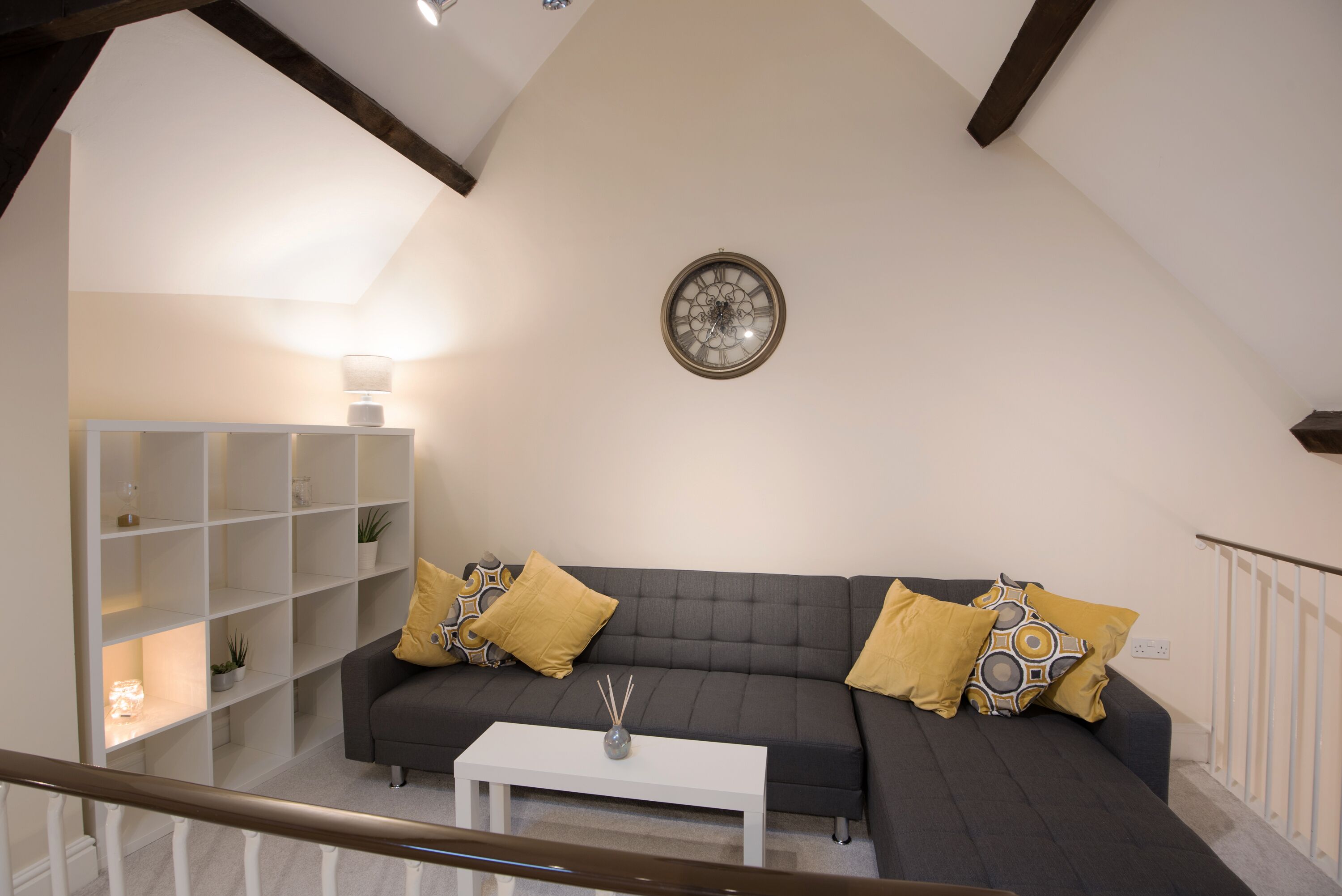 A modern living room with a dark grey sofa, white coffee table, and decorative pillows. A white shelving unit with plants is on the left, and a wall clock is above the sofa.