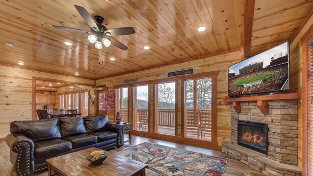 A cozy cabin interior with a leather sofa, a fireplace, a television, and large windows. - Mountain Moonshine, Private Indoor Pool, Pool Table, Arcade Game, Theater Room (Sevierville)