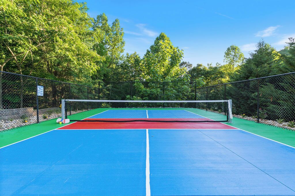 An outdoor tennis court with a net, surrounded by a chain-link fence and trees.
