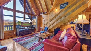 A spacious living room with a large window, a red leather sofa, a wooden coffee table, and a staircase leading to an upper level. - Tree Top. Beautiful Mountain View, Pool table, Hot Tub, Arcade Game, Private Mountain Setting (Sevierville)