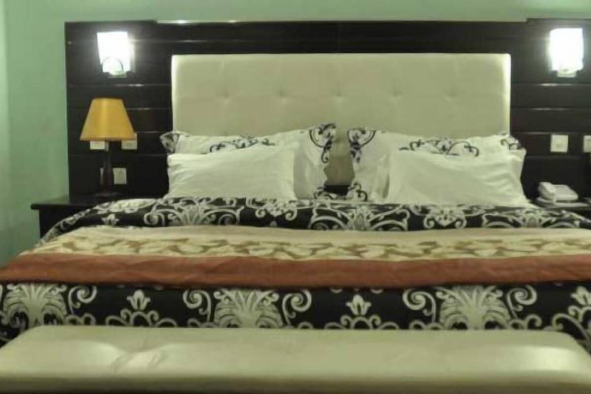 A neatly made bed with a patterned bedspread and pillows, a bedside lamp, and a nightstand with a phone and a cup.