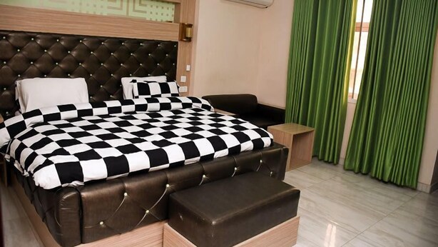 A bedroom with a checkered bedspread, a tufted headboard, a wooden nightstand, and green curtains. - Kelin Court Hotel (Lagos)
