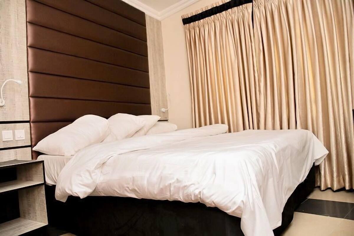 A hotel room with a bed, a wooden headboard, a nightstand, and beige curtains.