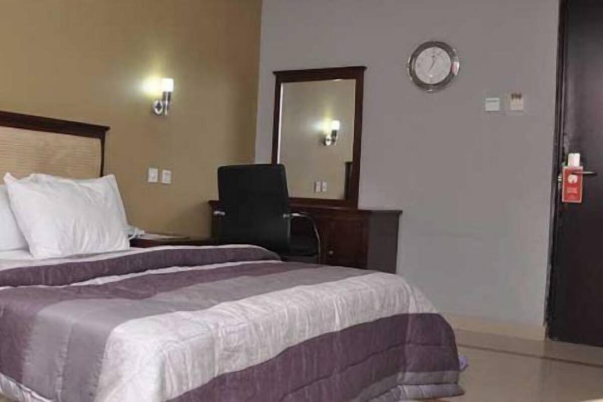 A hotel room with a bed, a chair, a desk, a mirror, and a clock on the wall.