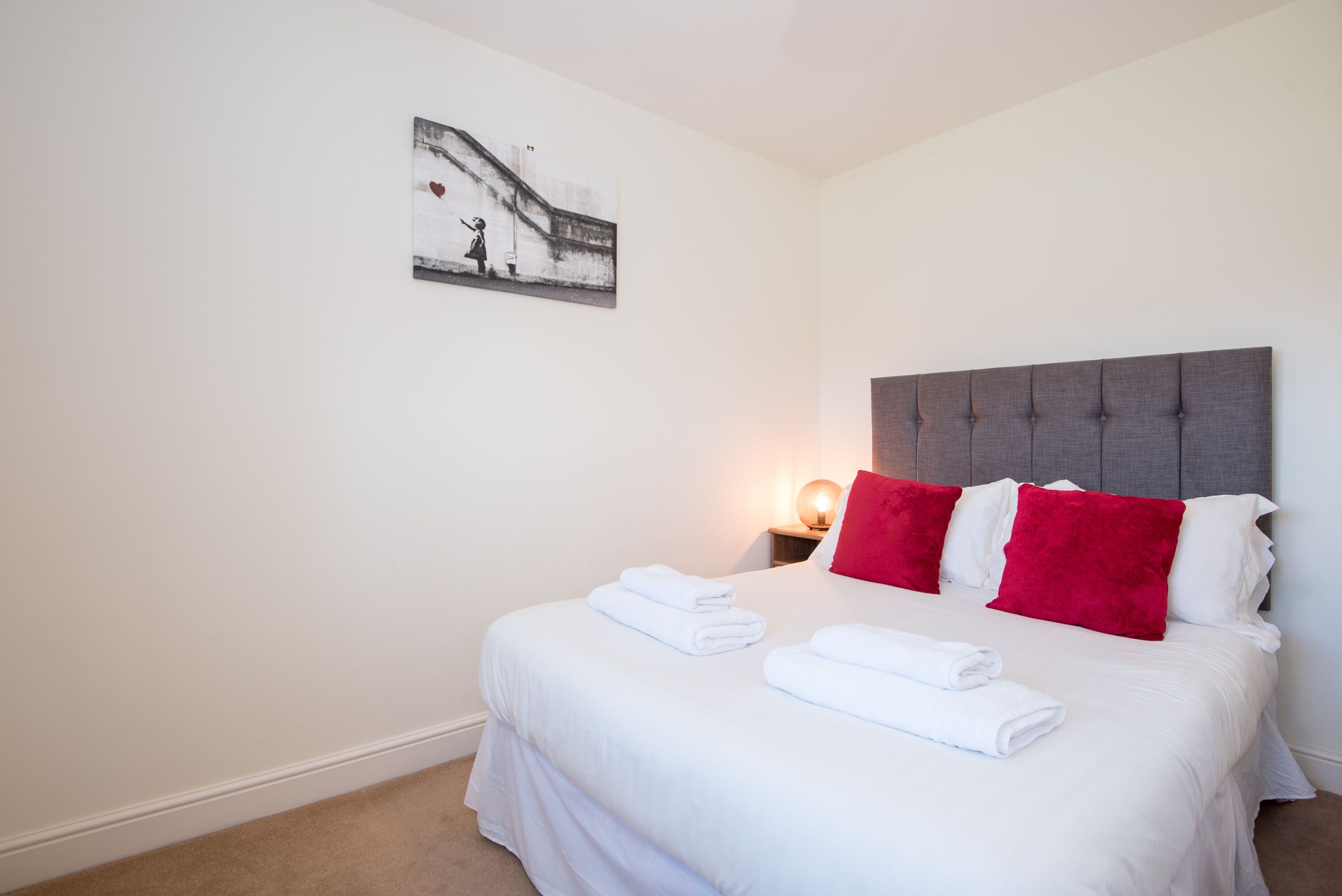 A neatly made bed with white and red pillows, a headboard, and a wall-mounted picture.