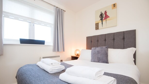 A neatly made bed with white and grey linens, a grey headboard, and a painting above it.