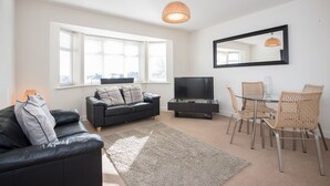 A modern living room with a black leather sofa, a glass coffee table, wicker chairs, a flat-screen TV, and a large window with blinds.