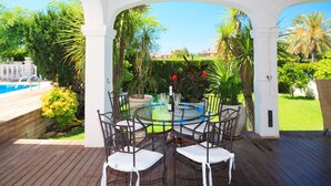 A covered outdoor patio with a glass table and chairs, surrounded by lush greenery and a view of a swimming pool. - Spacious villa with private pool and free wifi, sleeps 12 - VILLA NOSTRA (Cambrils)