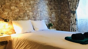 A bedroom with a stone wall, a bed with white bedding, a lamp, and a window with floral curtains. - Wine Inn Cascais (Cascais)