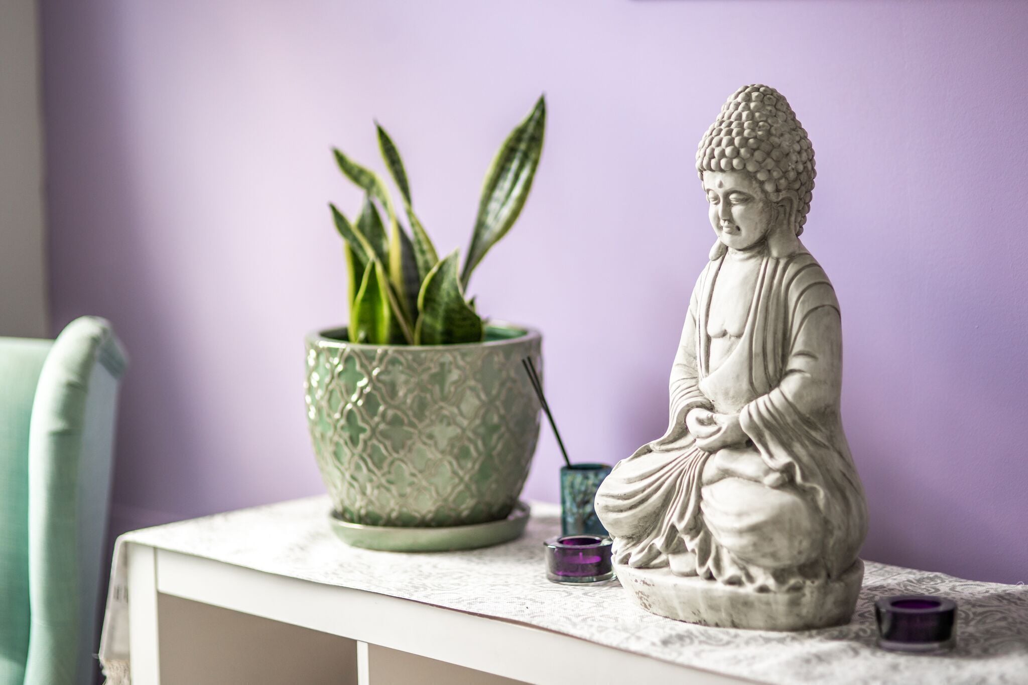 Eine Statue einer sitzenden Person, ein Blumentopf und zwei kleine Behälter auf einer weißen Oberfläche vor einer violetten Wand.