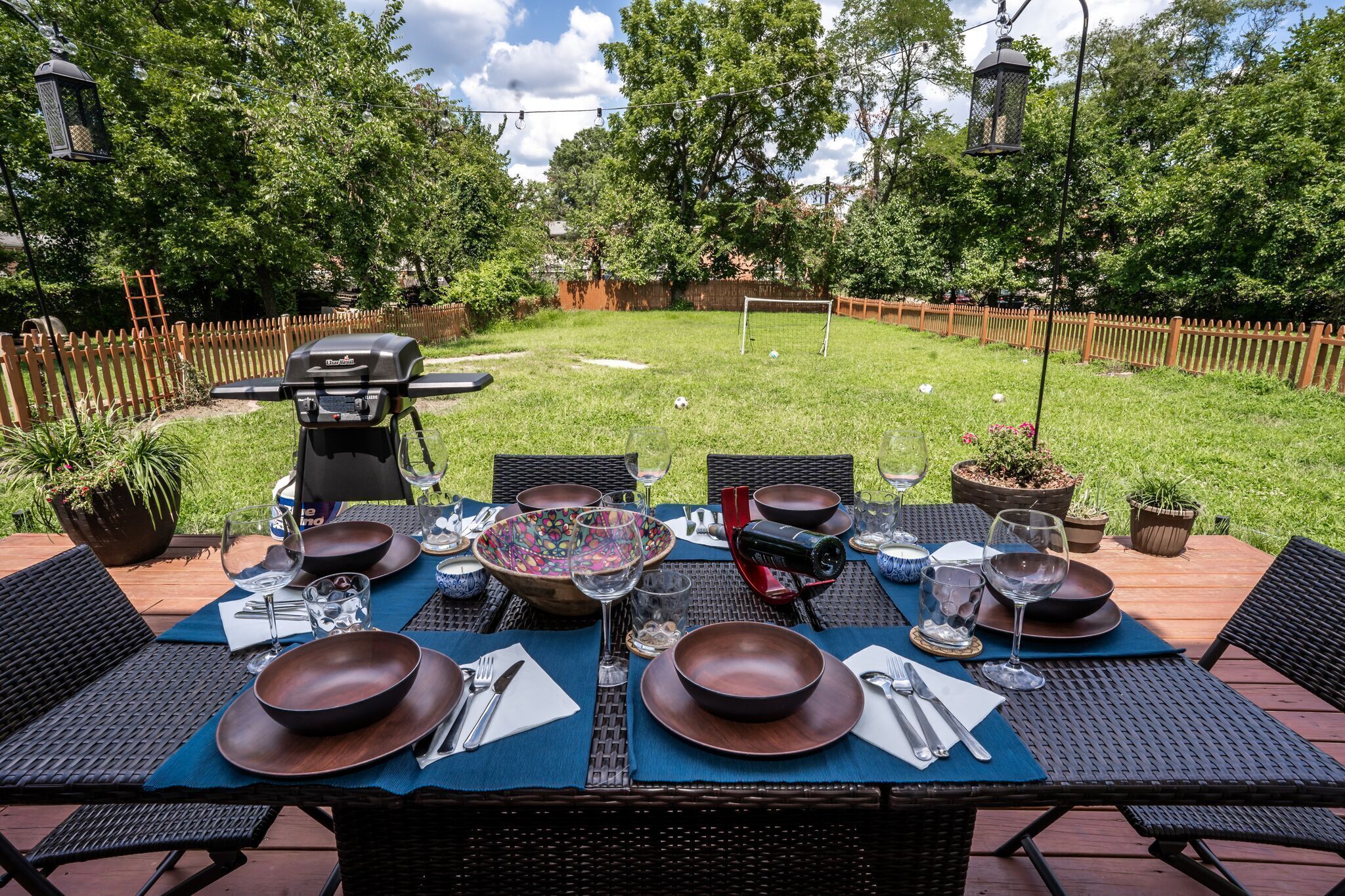 An outdoor dining area with a table set for four, surrounded by chairs, and a barbecue grill in the background.