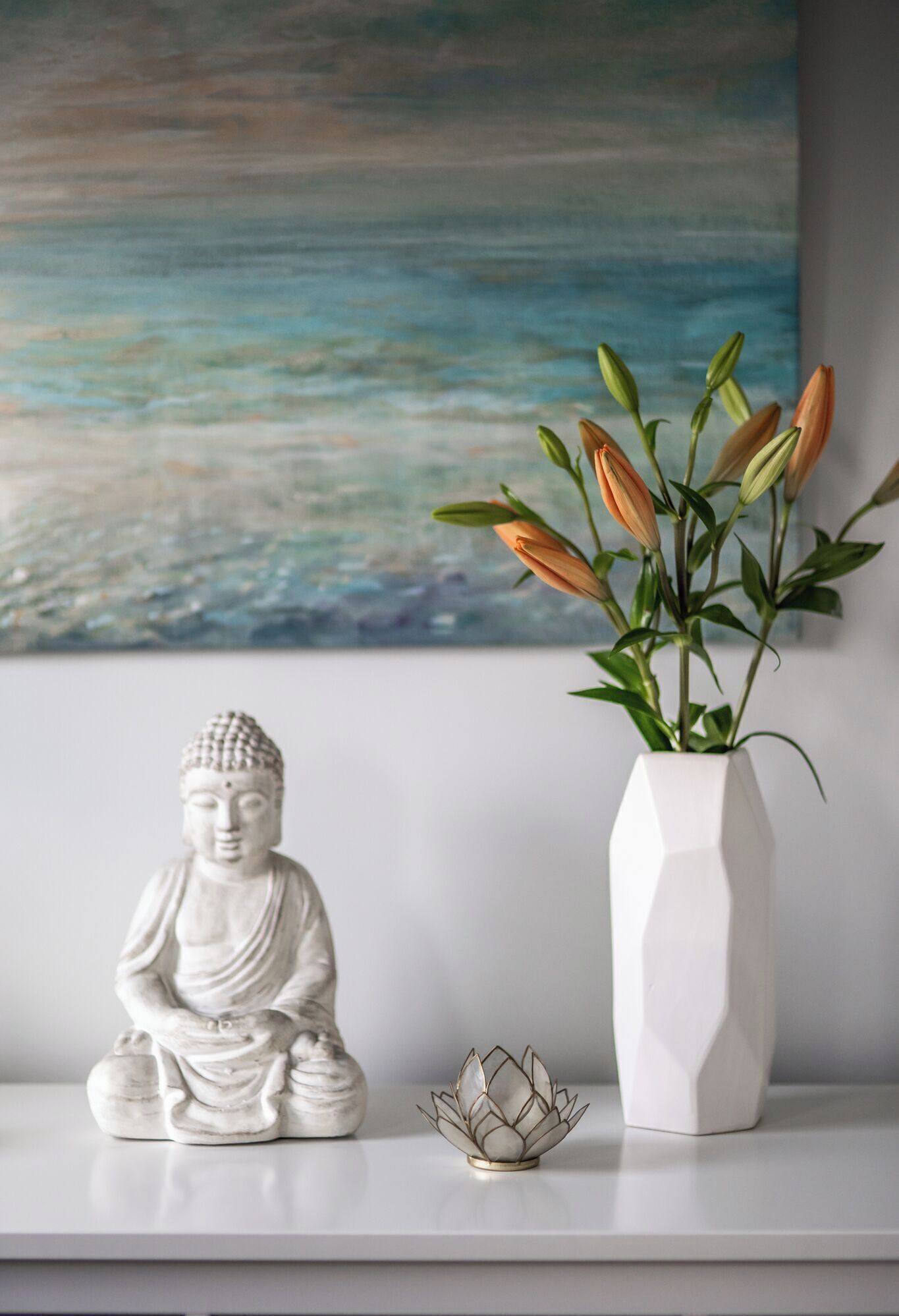 Een Boeddhabeeld, een witte vaas met bloemen en een schilderij op een witte plank.