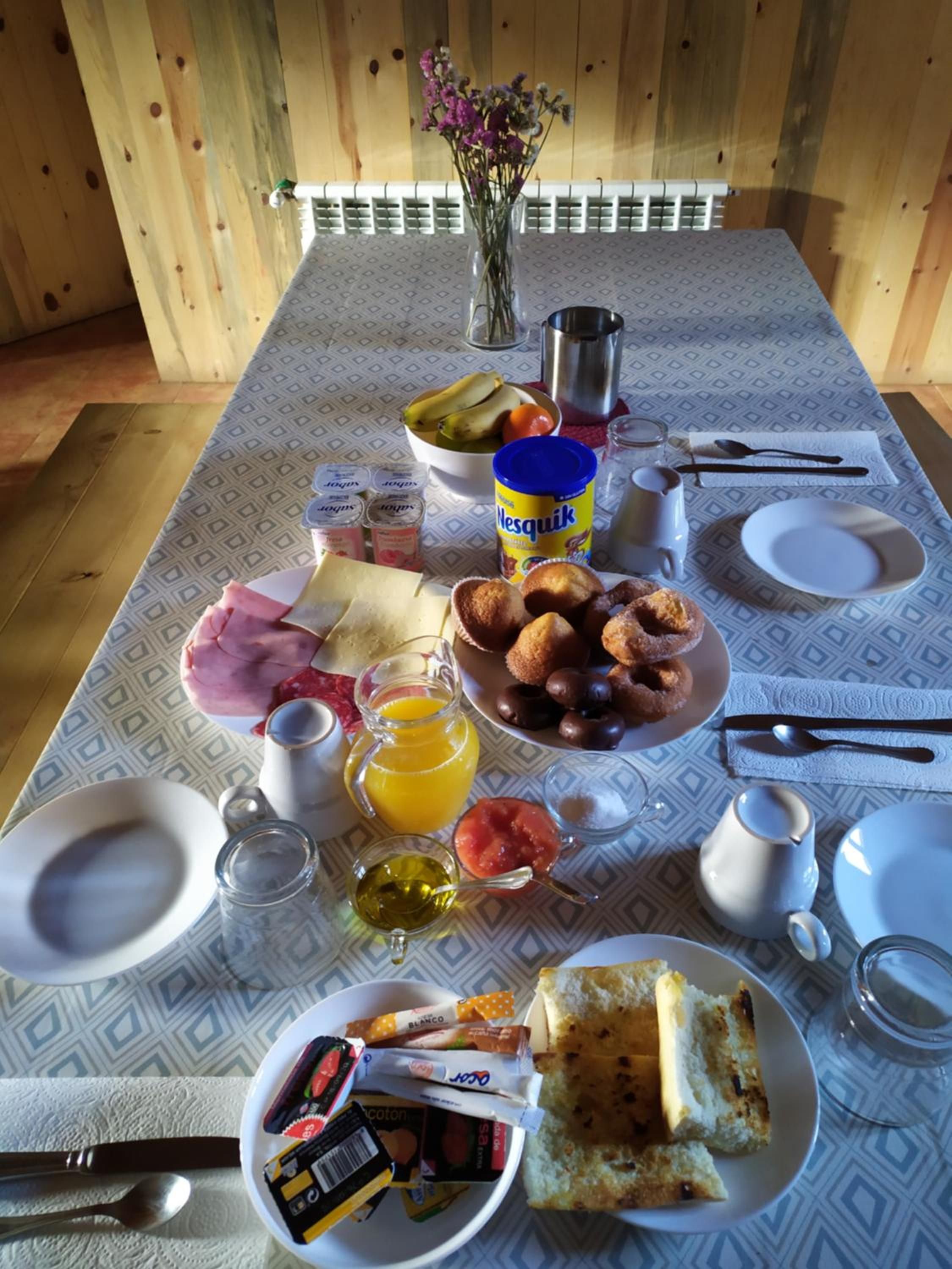 A breakfast table with various food items, a vase with flowers, and a patterned tablecloth.