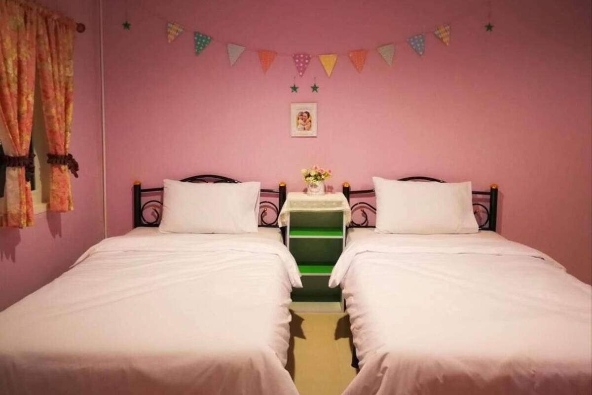 A twin room with two beds, a small table with a vase, and a pink wall with bunting.