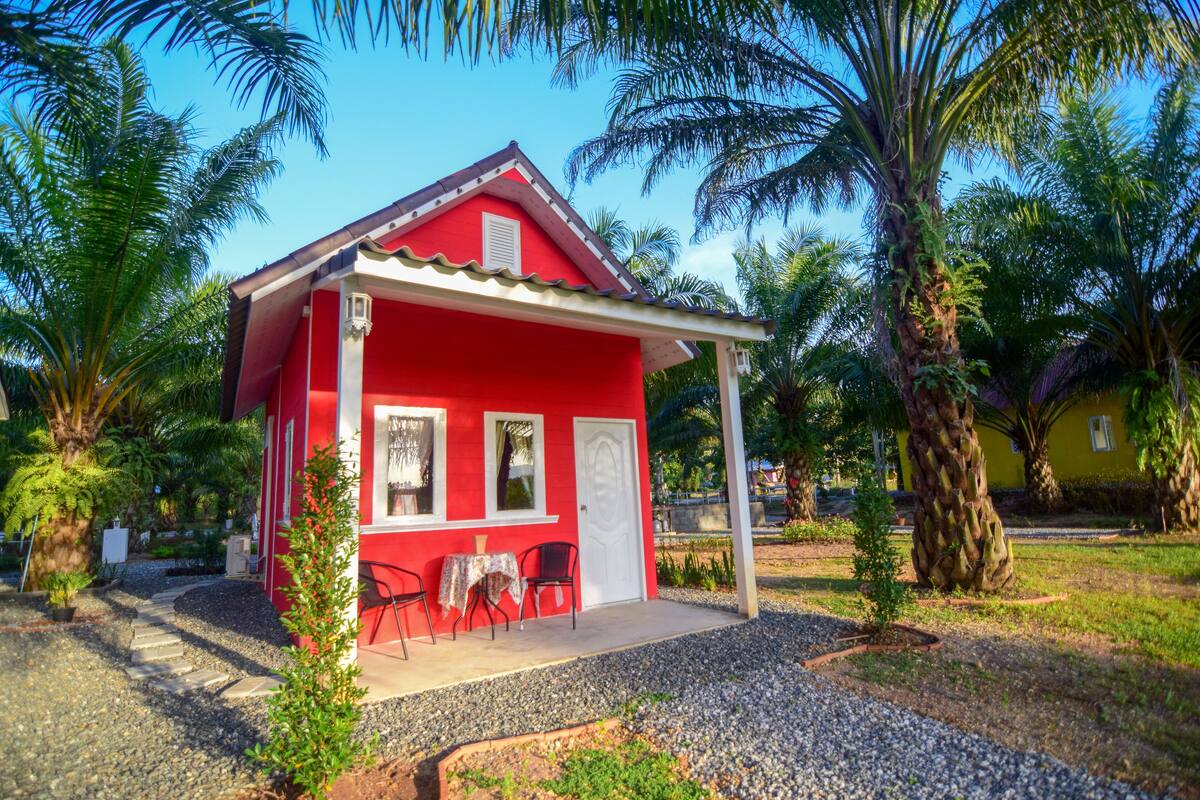 A red building with a white door and windows, surrounded by palm trees and a gravel path.