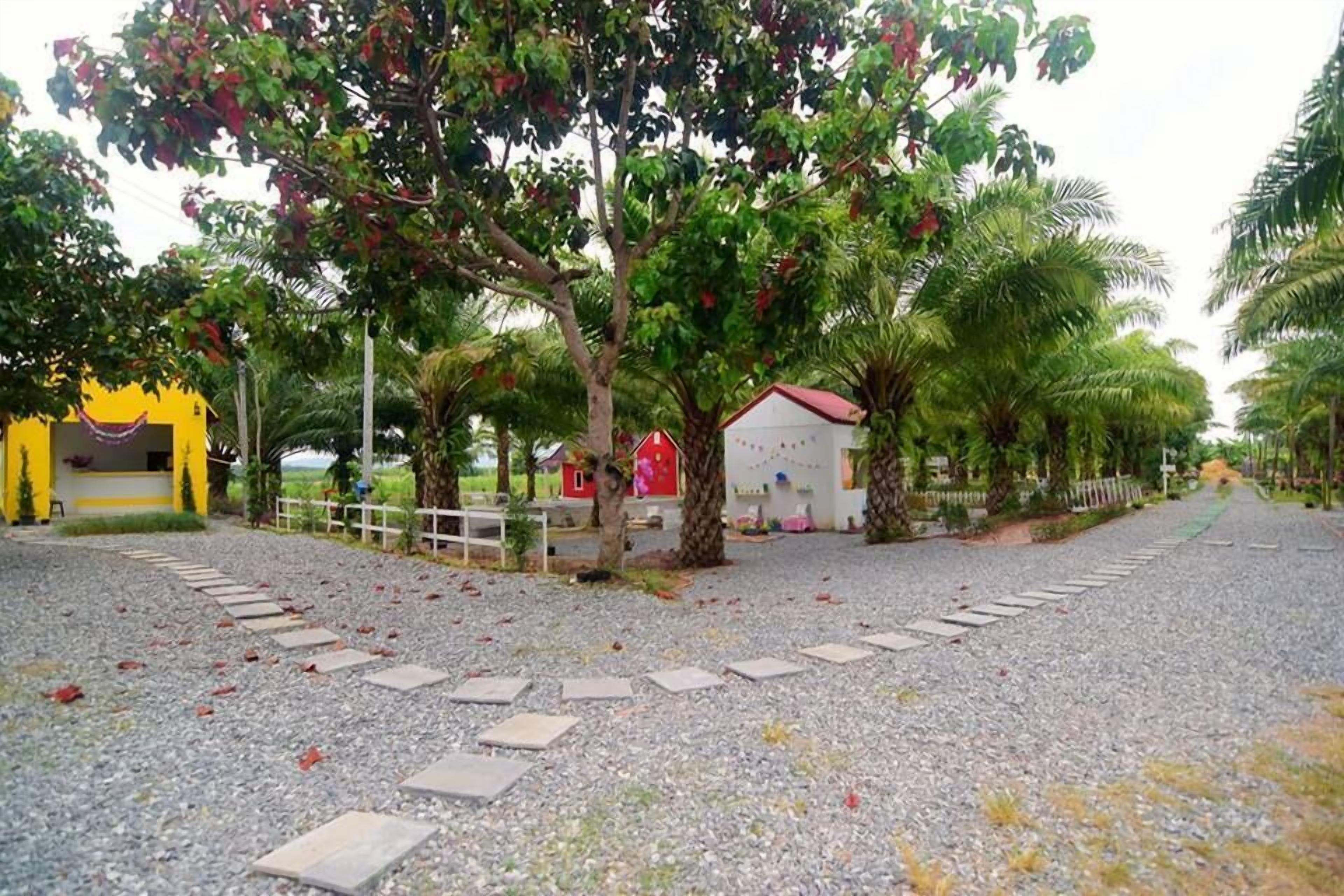 Un sendero de grava conduce a un pequeño edificio con techo rojo, rodeado de vegetación exuberante y palmeras.