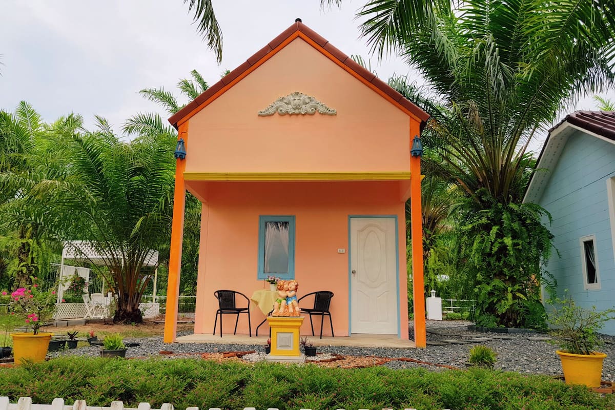 A small, single-story building with a peach-colored exterior and a white door. There are two black chairs and a small table in front of the door. The building is surrounded by lush greenery, including palm trees and various plants.