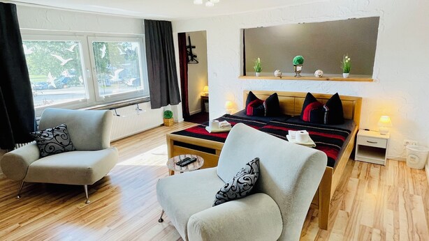 Ett modernt vardagsrum med en soffa, en fåtölj och en säng med ett rött och svart randigt överkast. Det finns ett fönster med gardiner, en växt på fönsterbrädan och en vägghylla med dekorativa föremål.