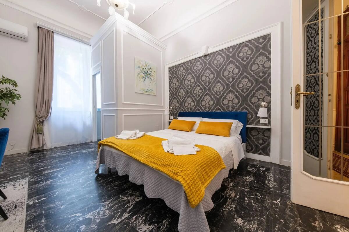 A hotel room with a bed, a blue chair, a plant, and a patterned wall.