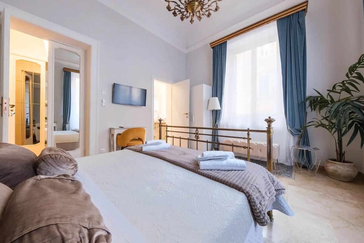 A well-lit bedroom with a large bed, a bedside table, a TV, and a potted plant.