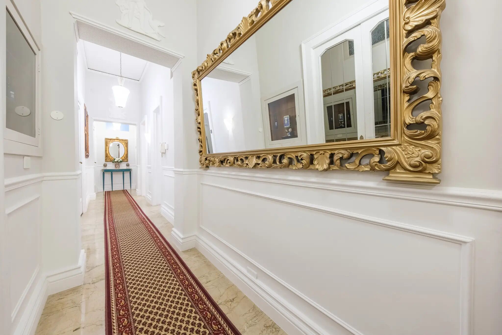Un pasillo con piso de mármol, alfombra roja estampada y un espejo grande con marco dorado ornamentado.