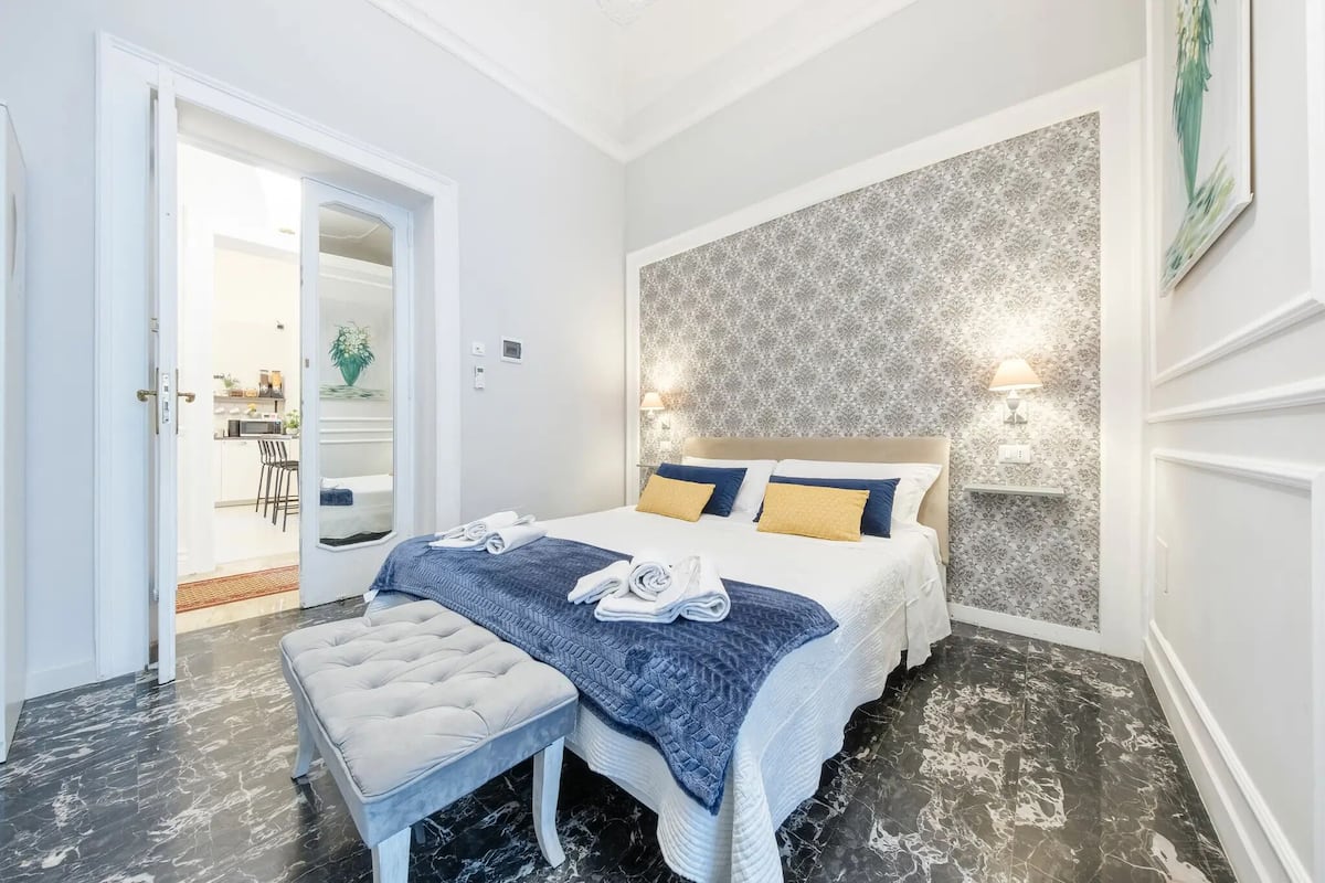 A bedroom with a bed, a bench, a patterned wall, and a door leading to a bathroom.