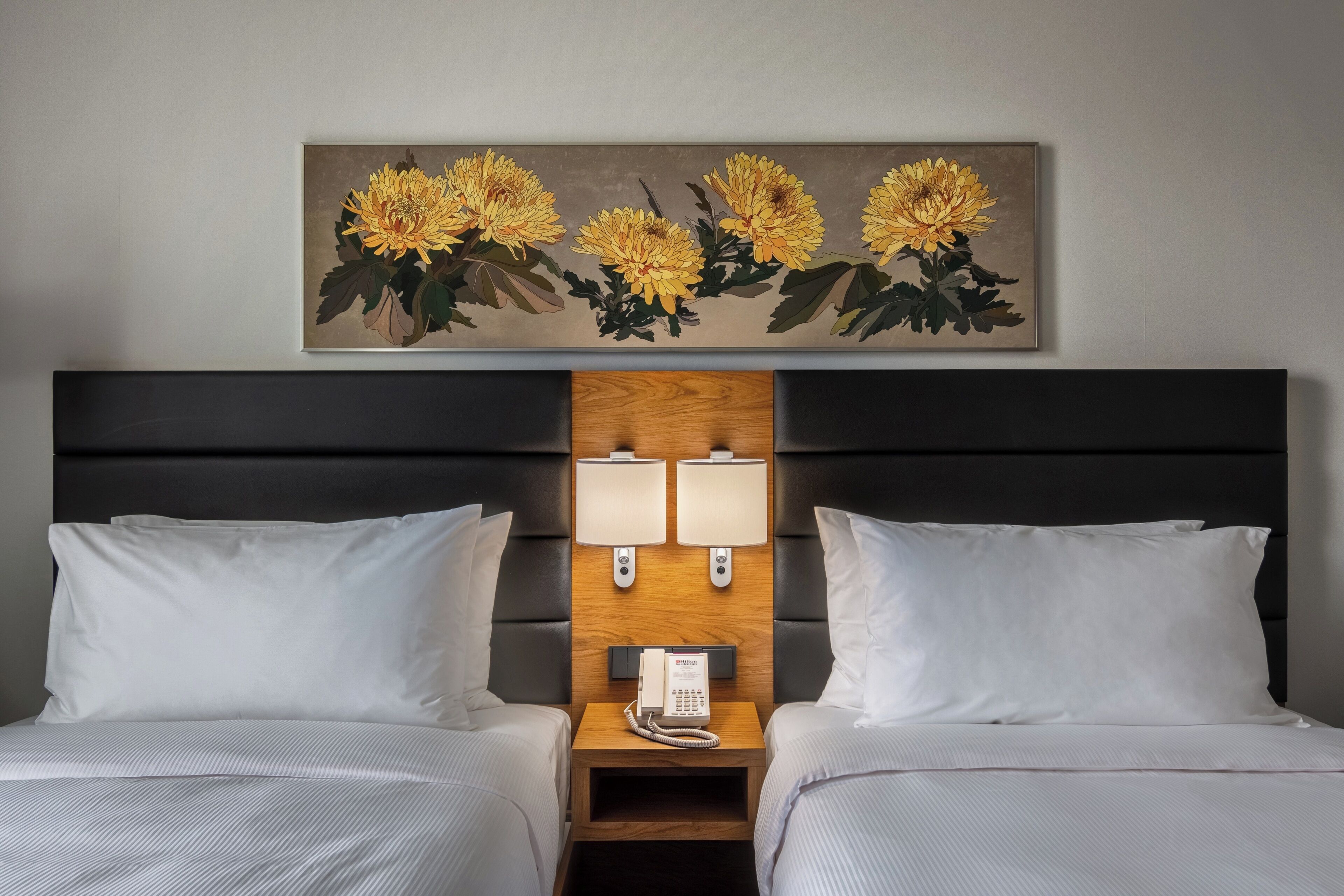 Habitación de hotel con dos camas, una mesita de noche con teléfono, dos lámparas de pared y un cuadro de flores amarillas.