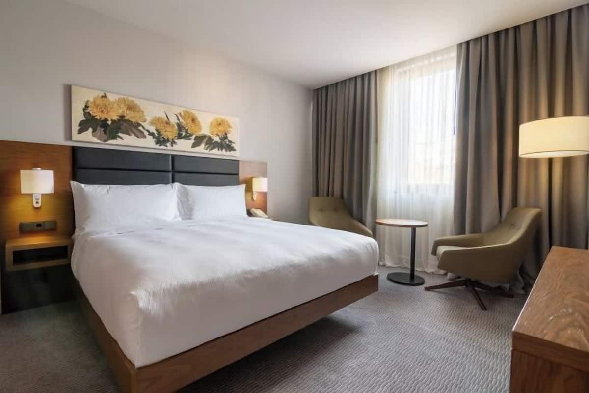 Een hotelkamer met een groot bed, een klein rond tafeltje, een stoel en een schilderij aan de muur.