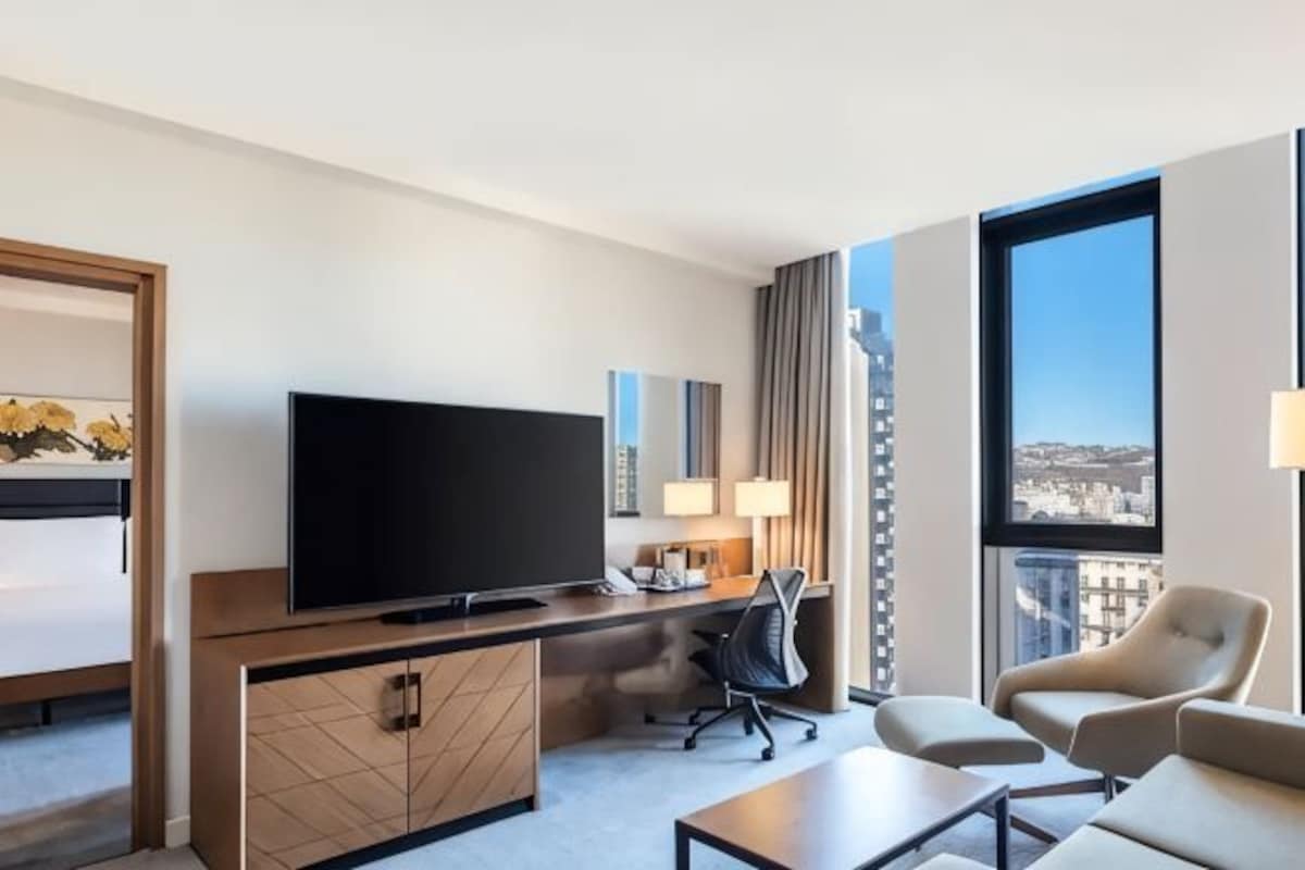 A hotel room with a flat-screen TV, a desk, a chair, a sofa, and a large window offering a city view.