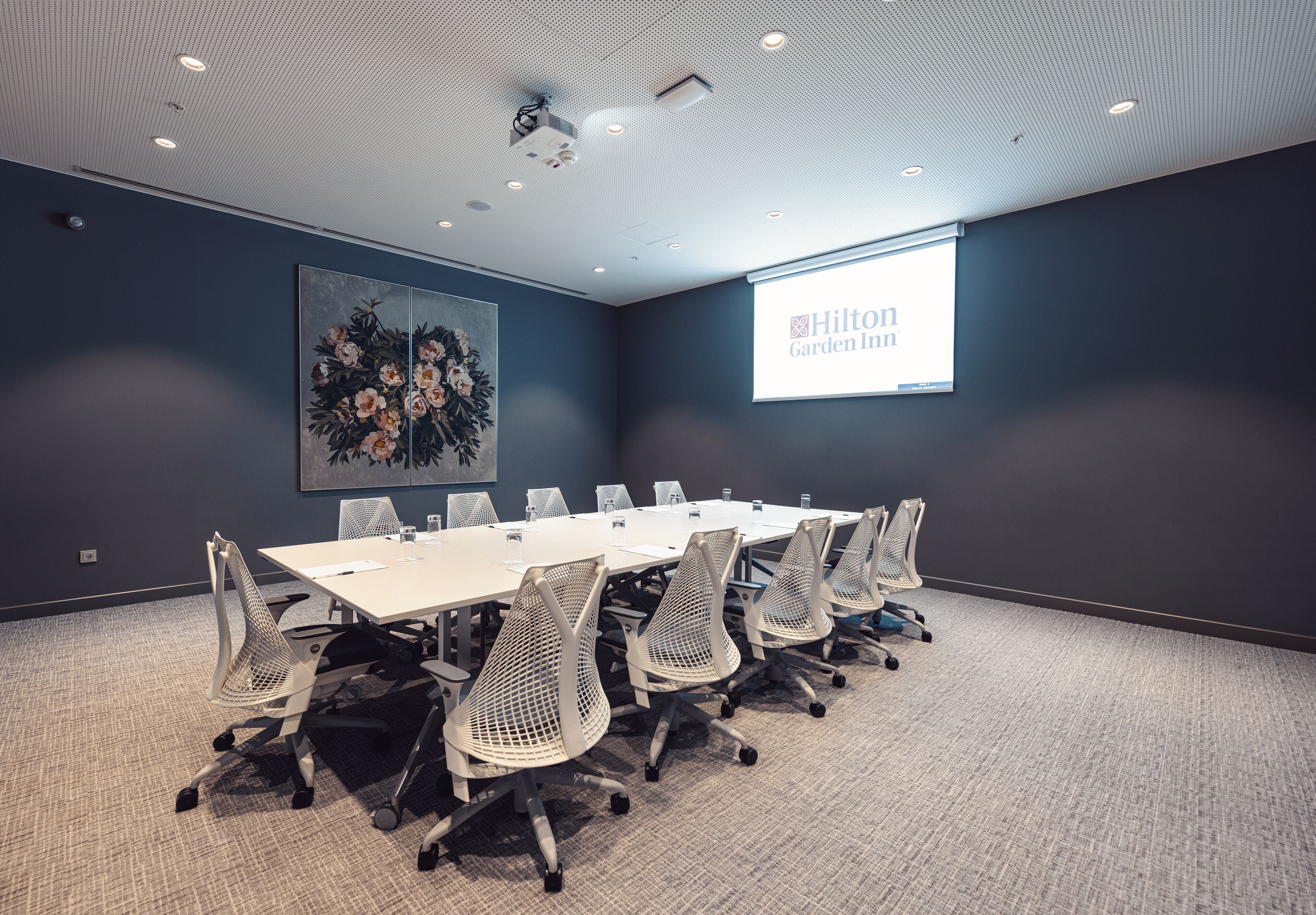 A conference room with a long table, chairs, a projector screen, and a floral painting on the wall.