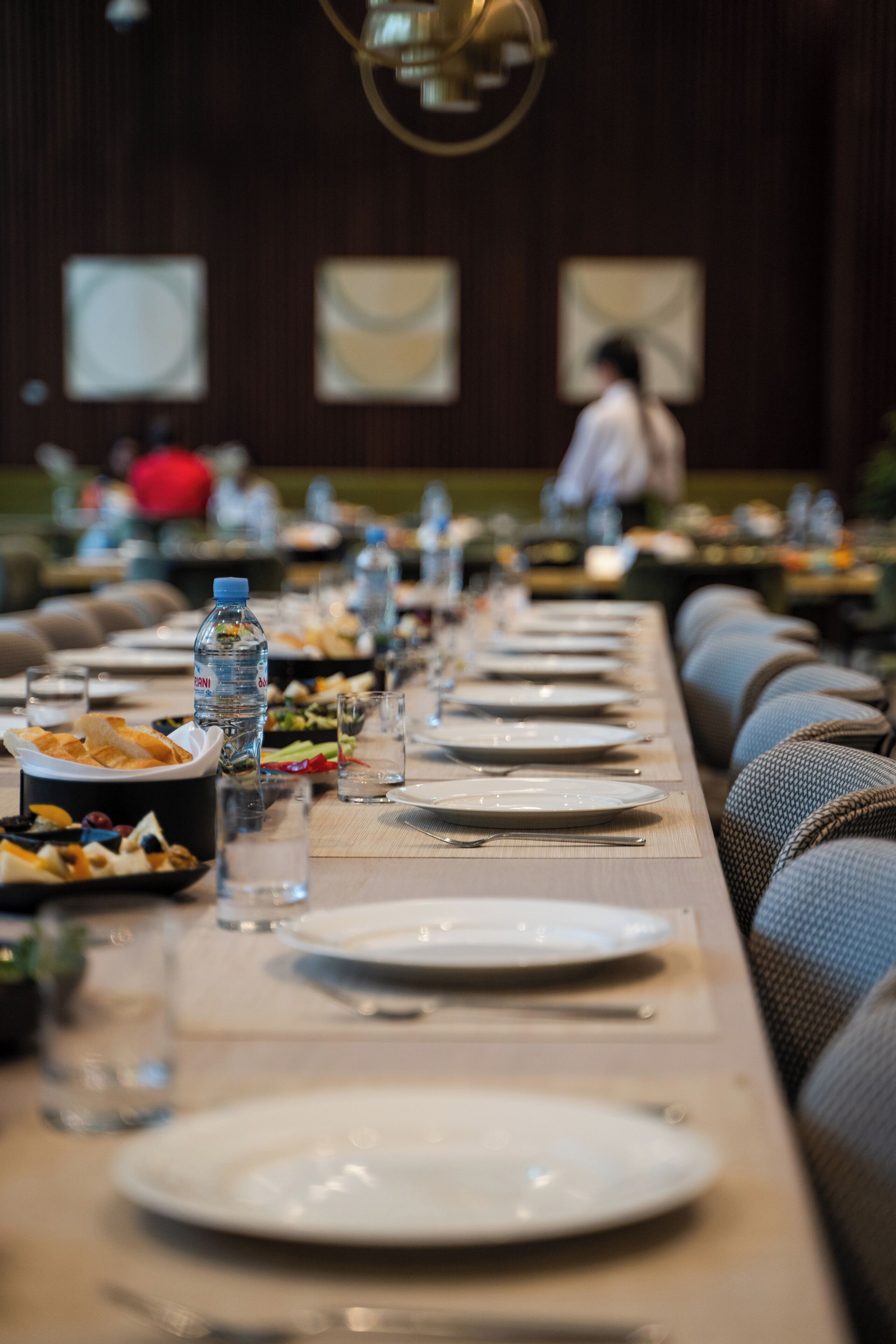 Una larga mesa de comedor con platos, vasos y botellas de agua, en una habitación con paredes de madera oscura y un candelabro.