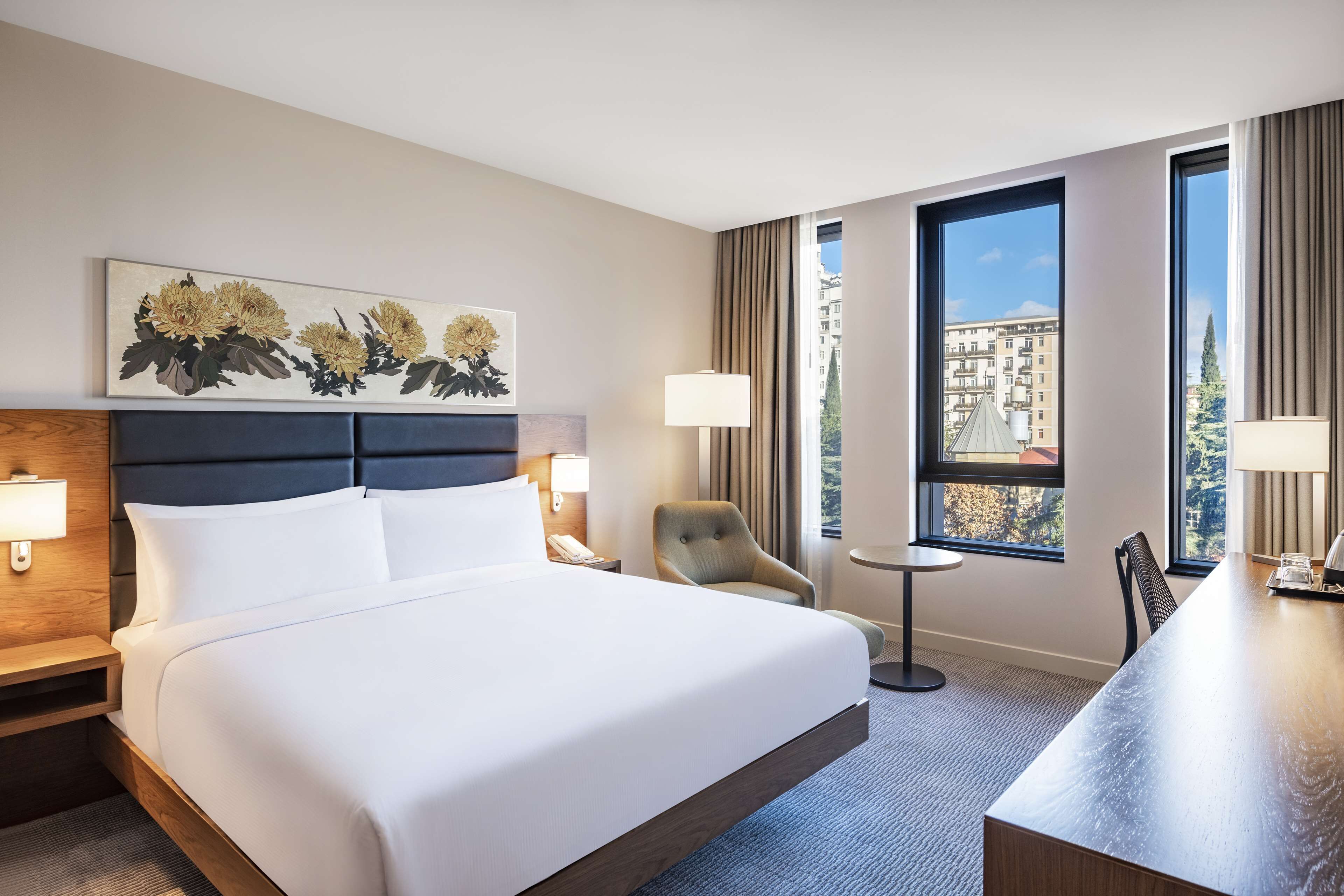 Ein Hotelzimmer mit einem großen Bett, einem Schreibtisch, einem Sessel und Blick auf Gebäude durch die Fenster.
