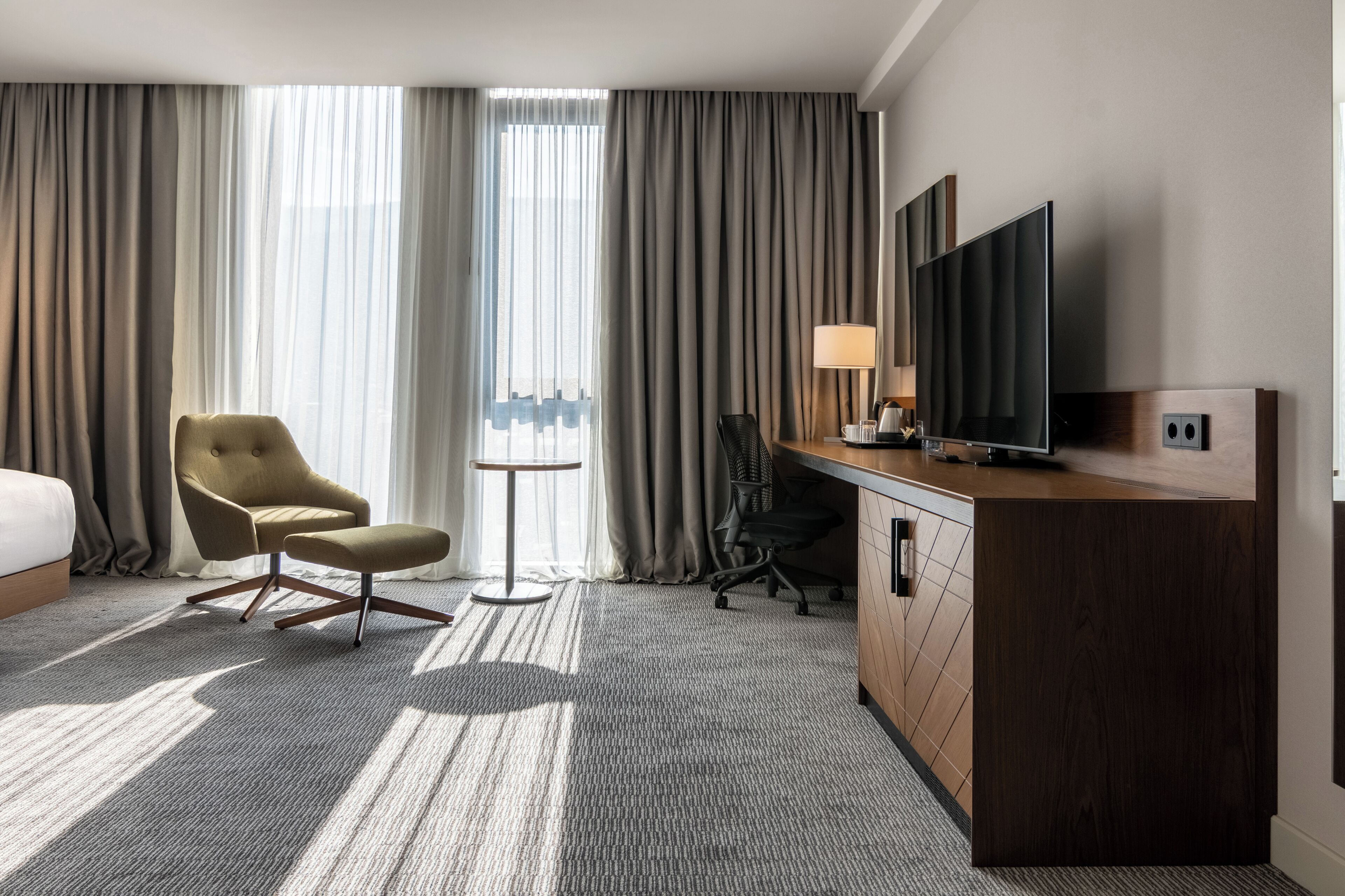A hotel room with a bed, a desk with a TV, a chair, a small table, and curtains.
