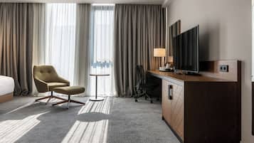 A hotel room with a bed, a desk with a TV, a chair, a small table, and curtains.