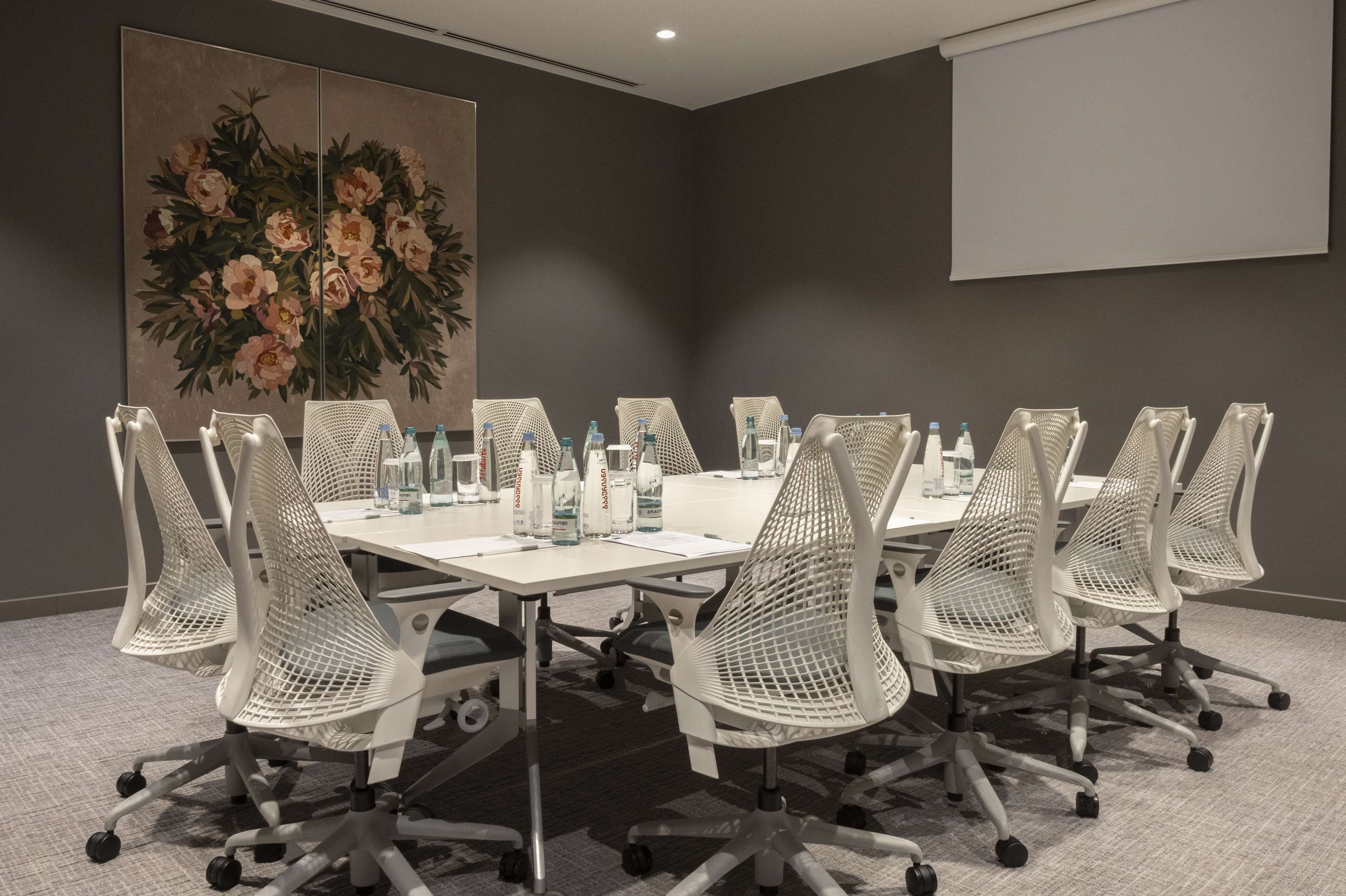 Ein Konferenzraum mit einem langen Tisch, weißen Mesh-Stühlen, Wasserflaschen und einem großen floralen Gemälde an der Wand.