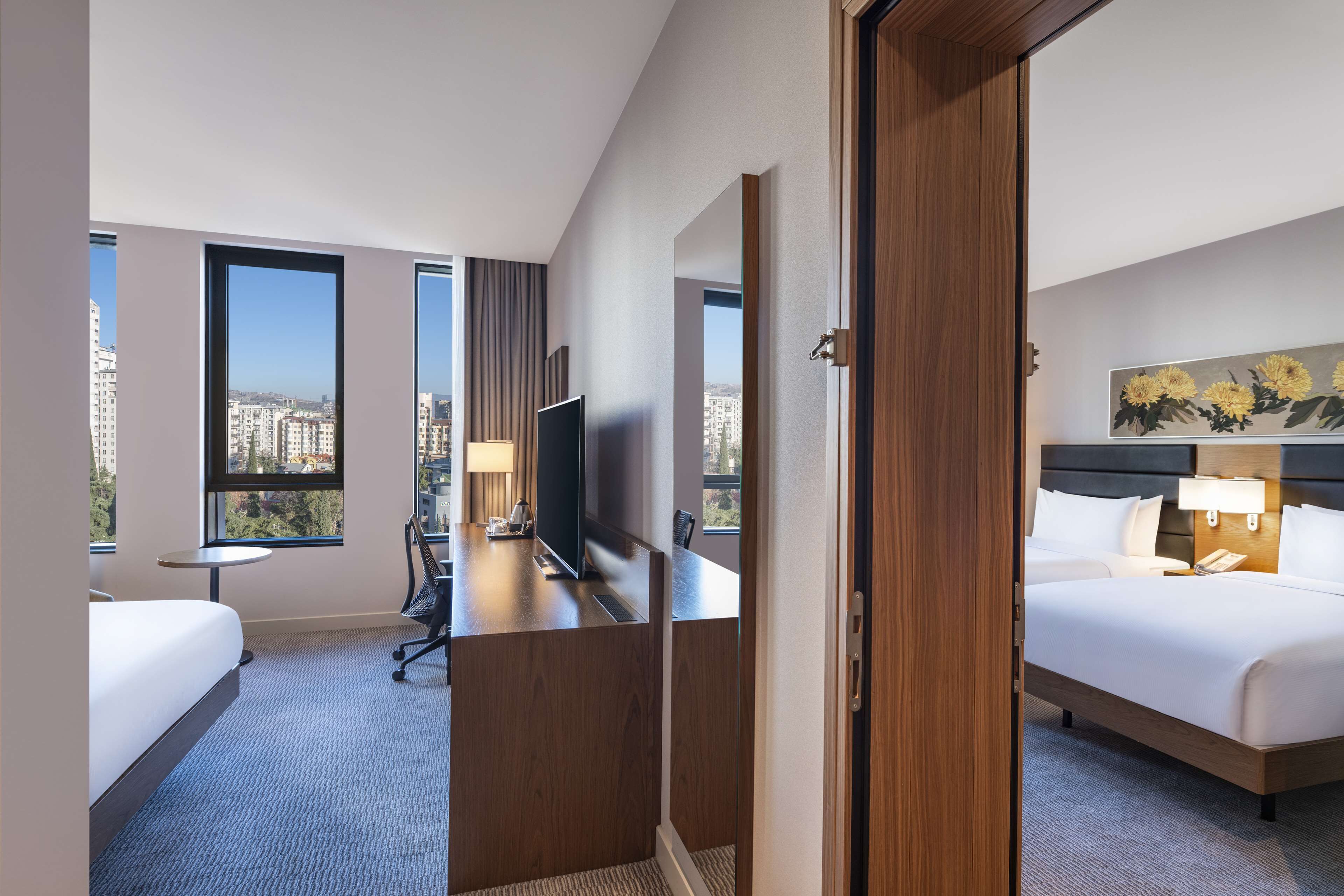 A hotel room with a bed, desk, chair, and a TV. There are large windows with a city view.