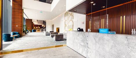 A modern hotel lobby with a marble reception desk, seating areas, and a staircase leading to an upper level.